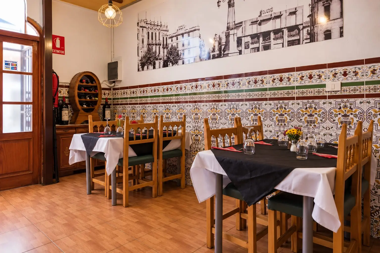 Comedor con las mesas puestas: mantel negro, servilletas rojas y flores, delante de una pared de azulejos valencianos.