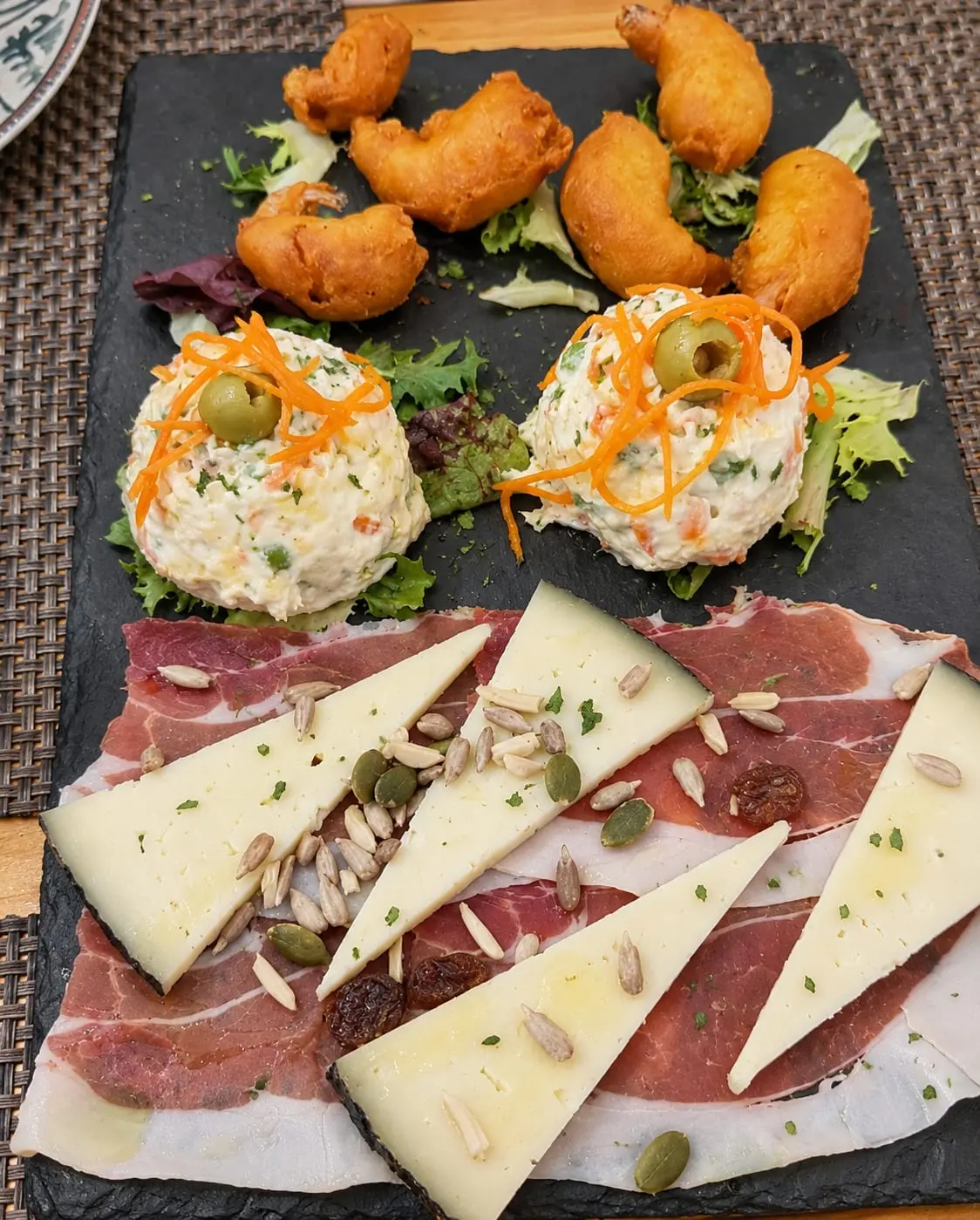 Tabla de pizarra para compartir: buñuelos fritos, dos raciones de ensaladilla y una tabla de jamón con cuñas de queso.