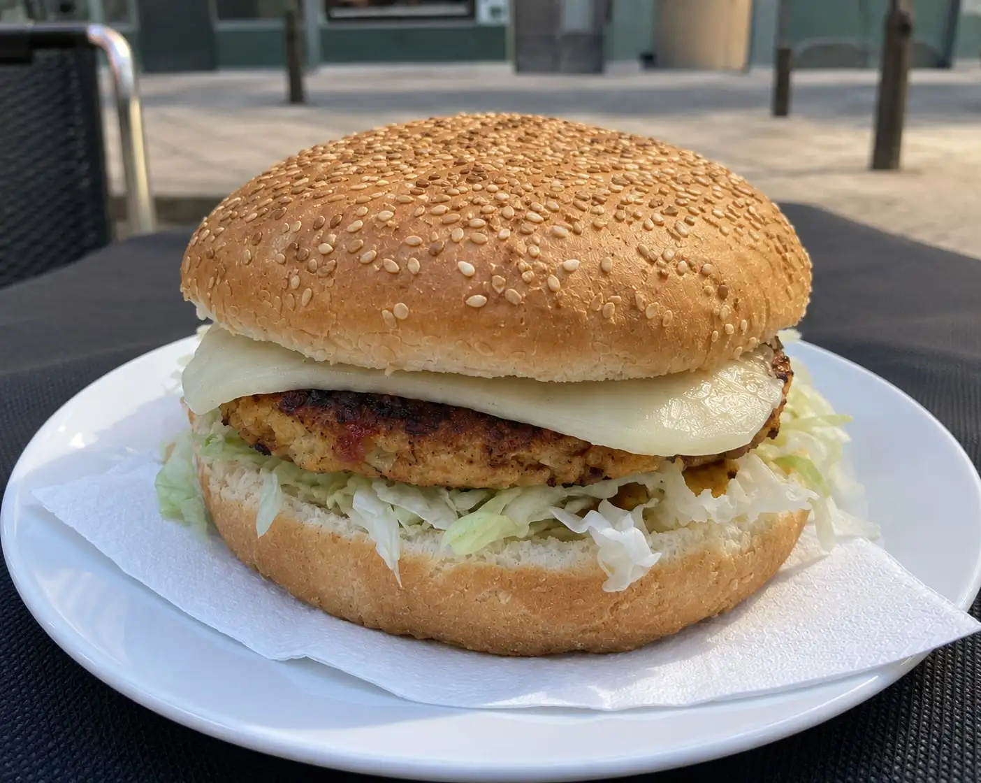 Hamburguesa en pan de sésamo con queso fundido y lechuga, servida en un plato blanco en la terraza.