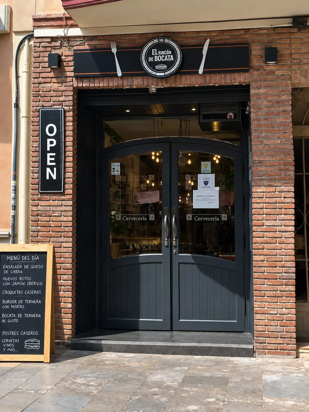 Entrada de El Rincón del Bocata: puerta doble oscura entre pilares de ladrillo, rótulo con cubiertos y pizarra con el menú del día en la acera.