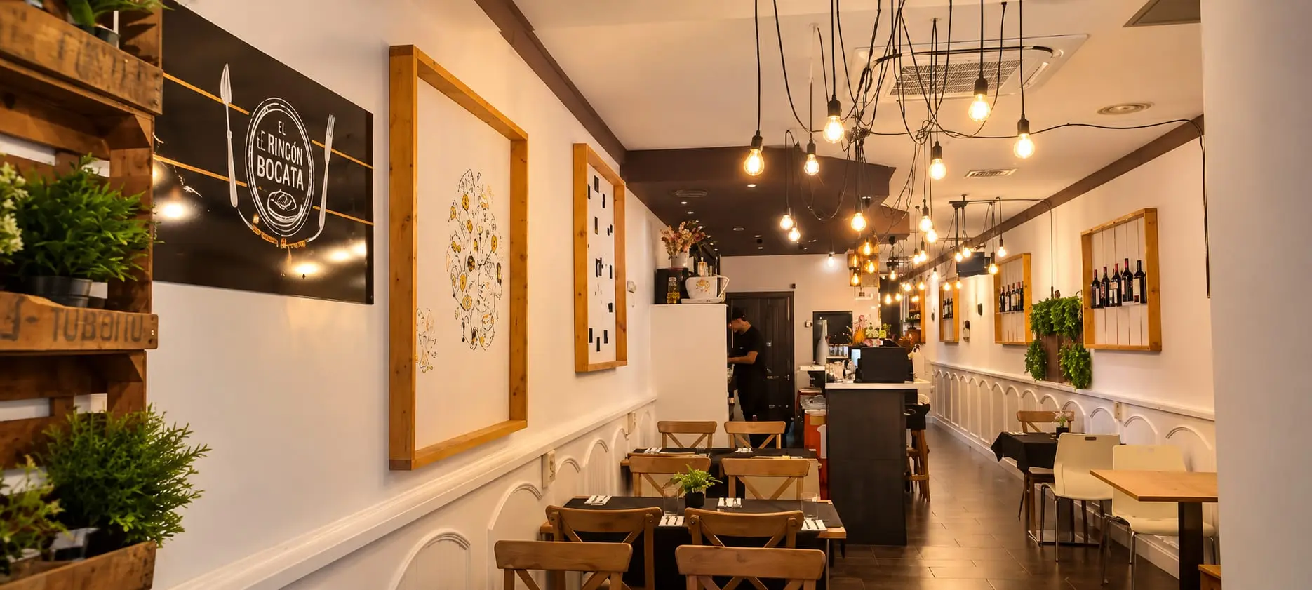 La sala alargada del restaurante: mesas de madera a un lado, la barra al fondo y una hilera de bombillas colgando del techo.