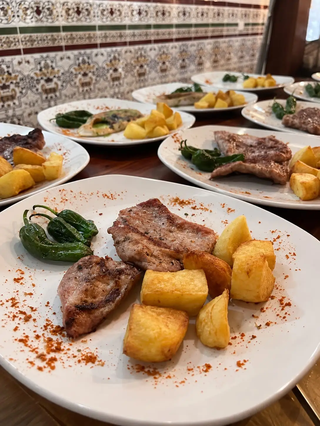 Varios platos servidos en fila sobre la mesa: carne a la plancha con patatas y pimientos verdes, delante de la pared de azulejos del comedor.