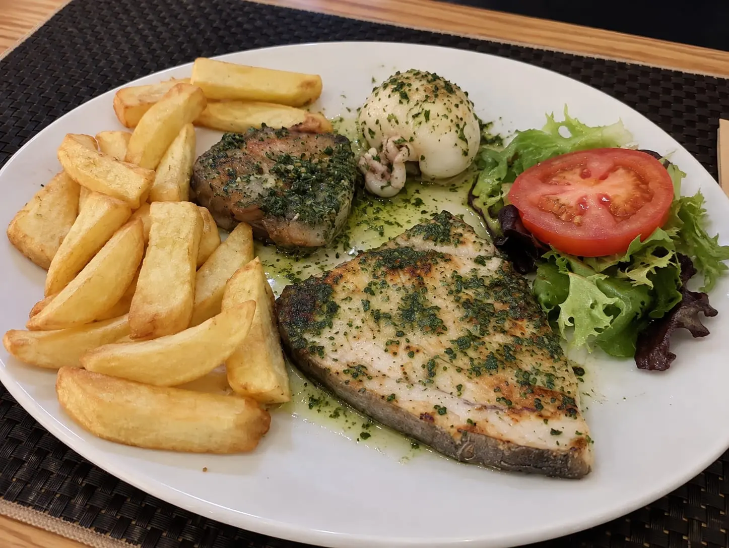 Plato de pescado a la plancha con salsa verde, un calamar relleno, patatas fritas y ensalada con tomate.