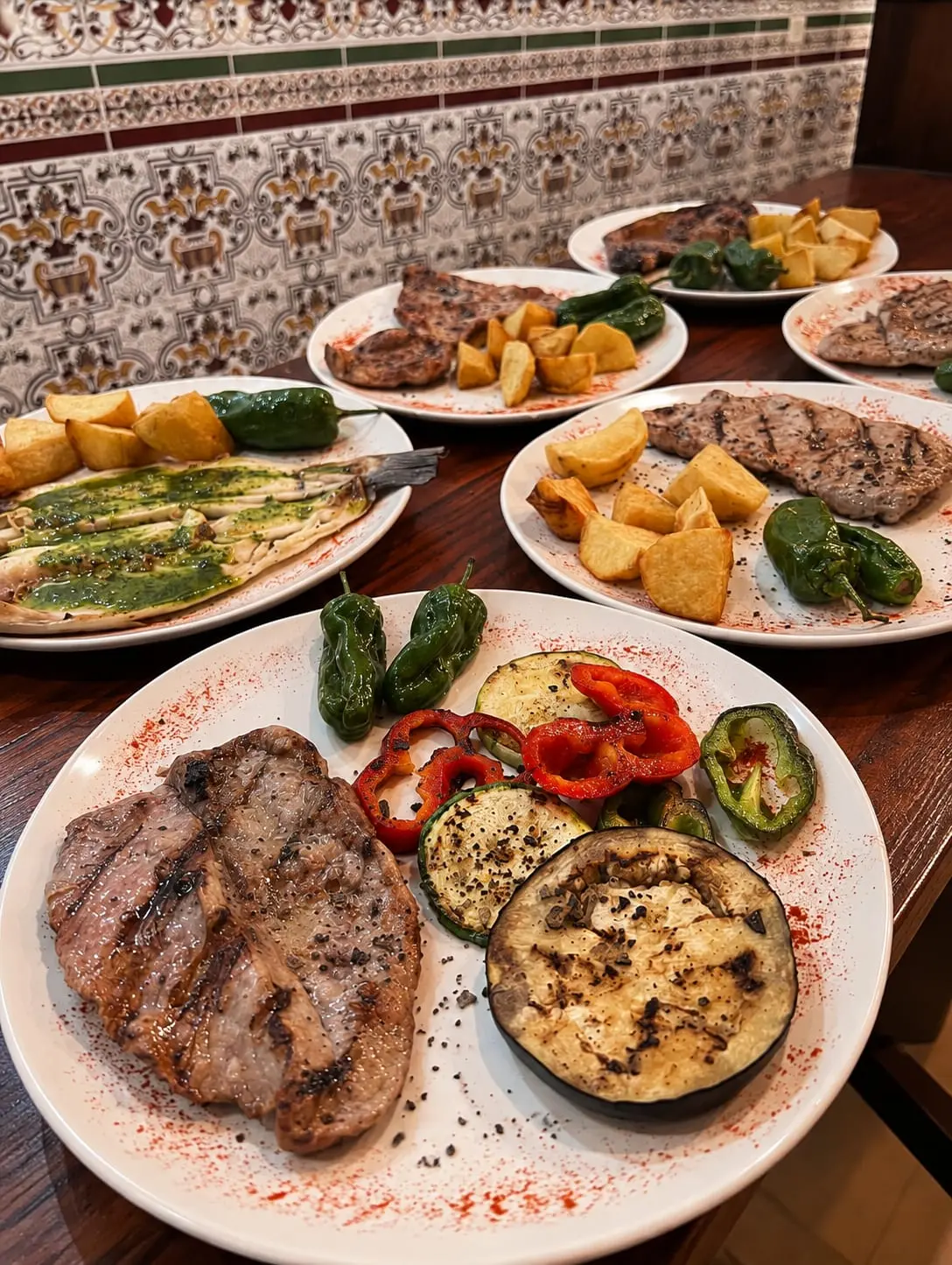 Mesa con varios platos: filete a la plancha con verduras asadas, y al fondo una fuente con pescado y salsa verde.