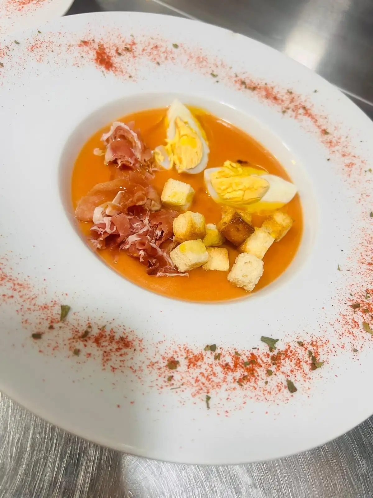 Salmorejo en plato hondo con huevo duro picado, virutas de jamón serrano y picatostes, con pimentón espolvoreado en el borde.
