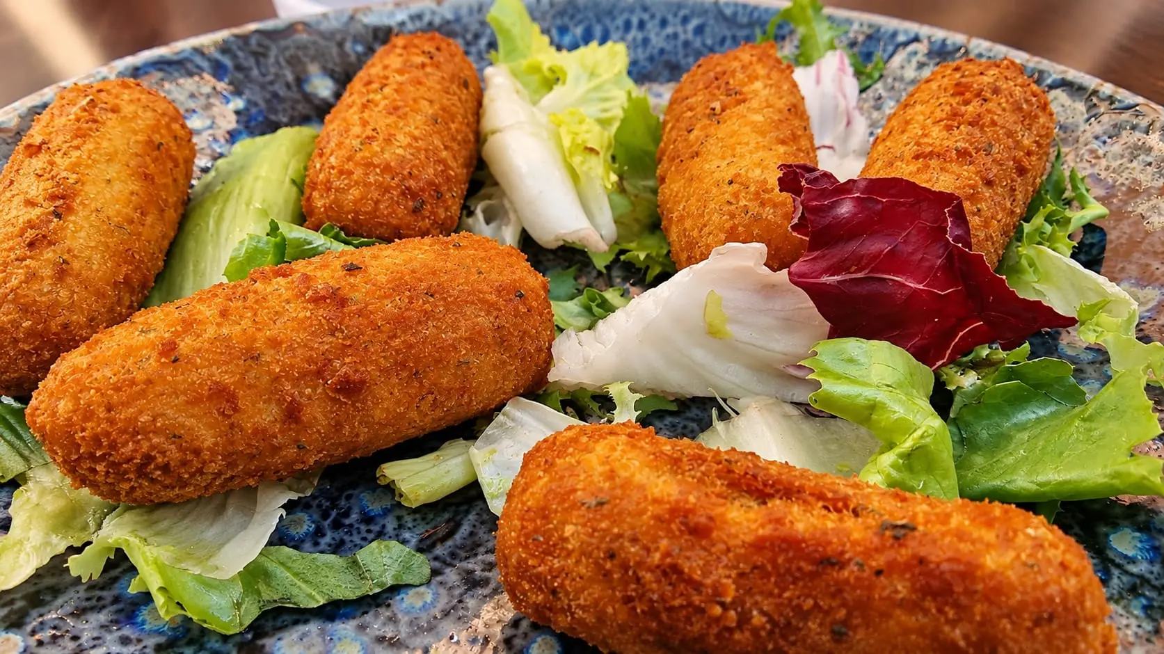 Seis croquetas recién fritas sobre un plato azul, con hojas de ensalada entre ellas.
