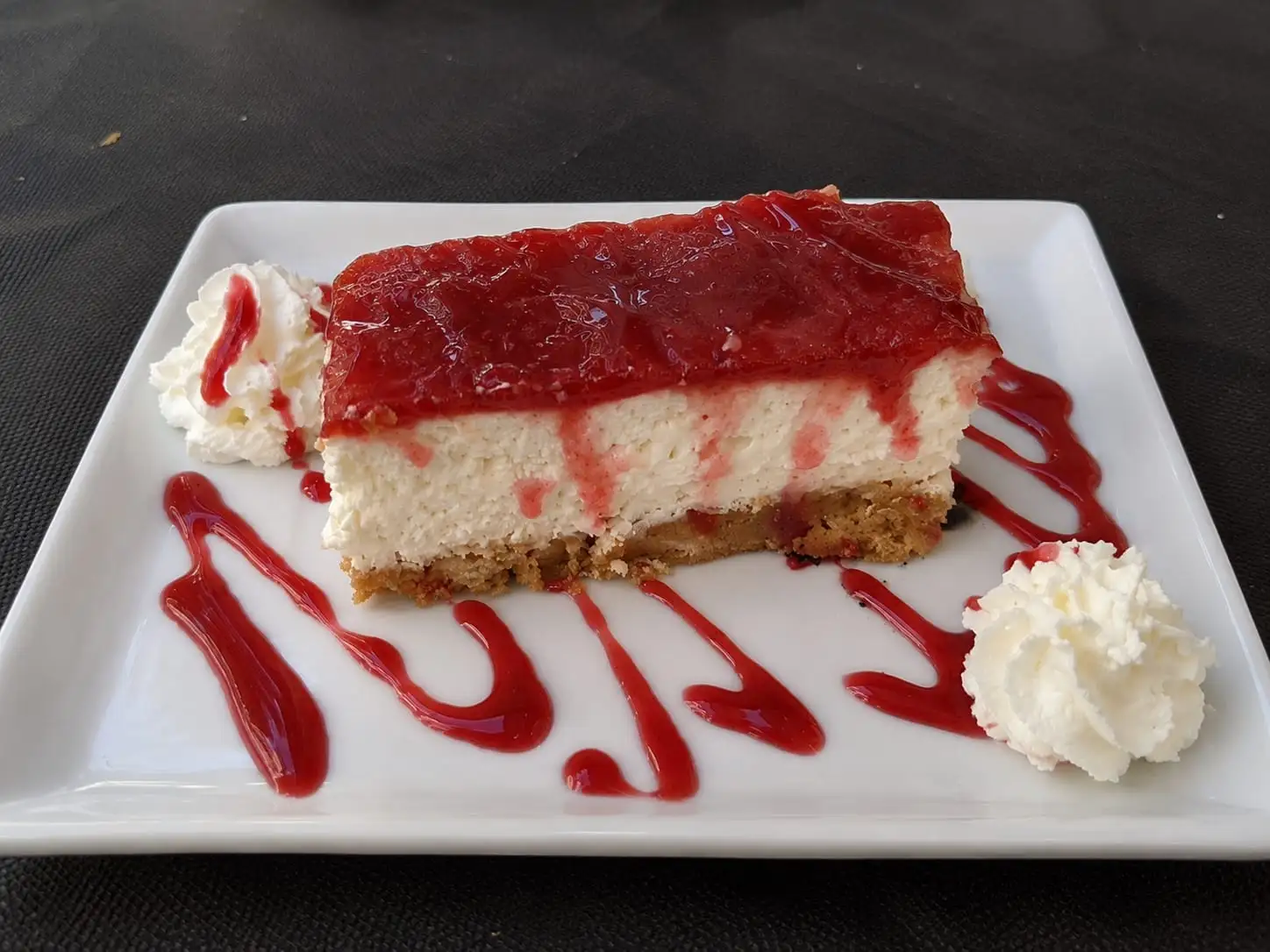 Ración de tarta de queso con mermelada de fresa por encima, nata montada a los lados y un hilo de coulis en el plato.