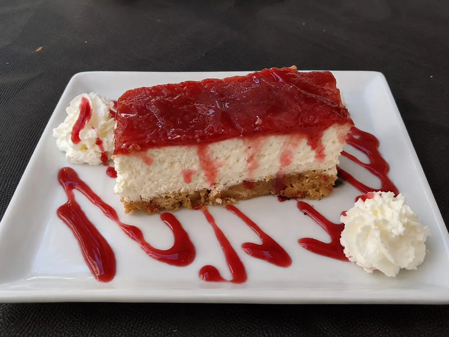 Ración de tarta de queso con mermelada de fresa por encima, nata montada a los lados y un hilo de coulis en el plato.