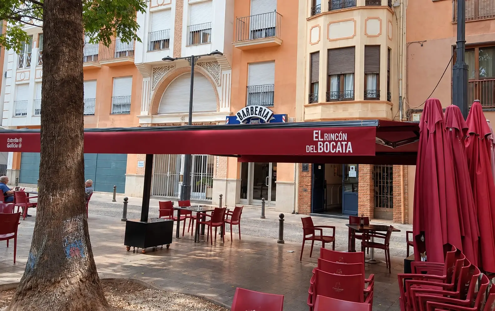 La terraza vista desde la plaza: el toldo granate con el nombre del restaurante sobre las mesas y las sillas rojas, a la sombra de un árbol.