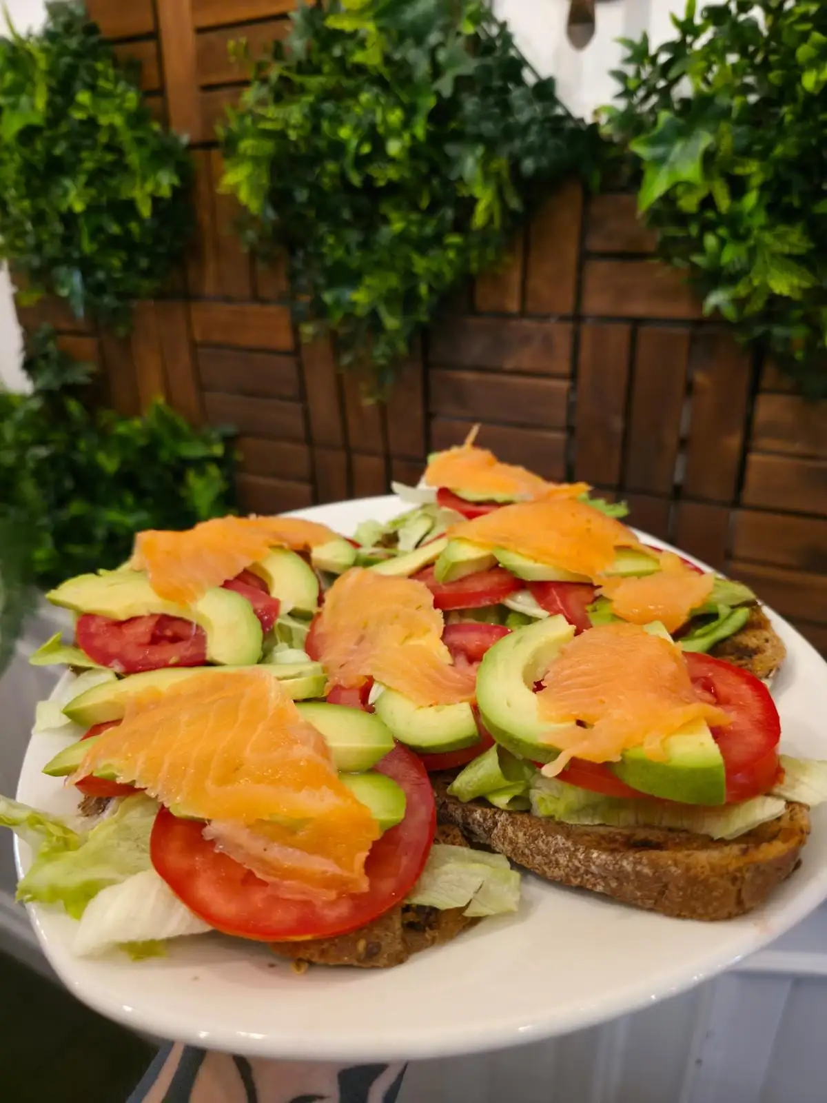 Dos tostadas de pan de cereales con lechuga, tomate, aguacate en láminas y salmón ahumado, servidas en un plato blanco.