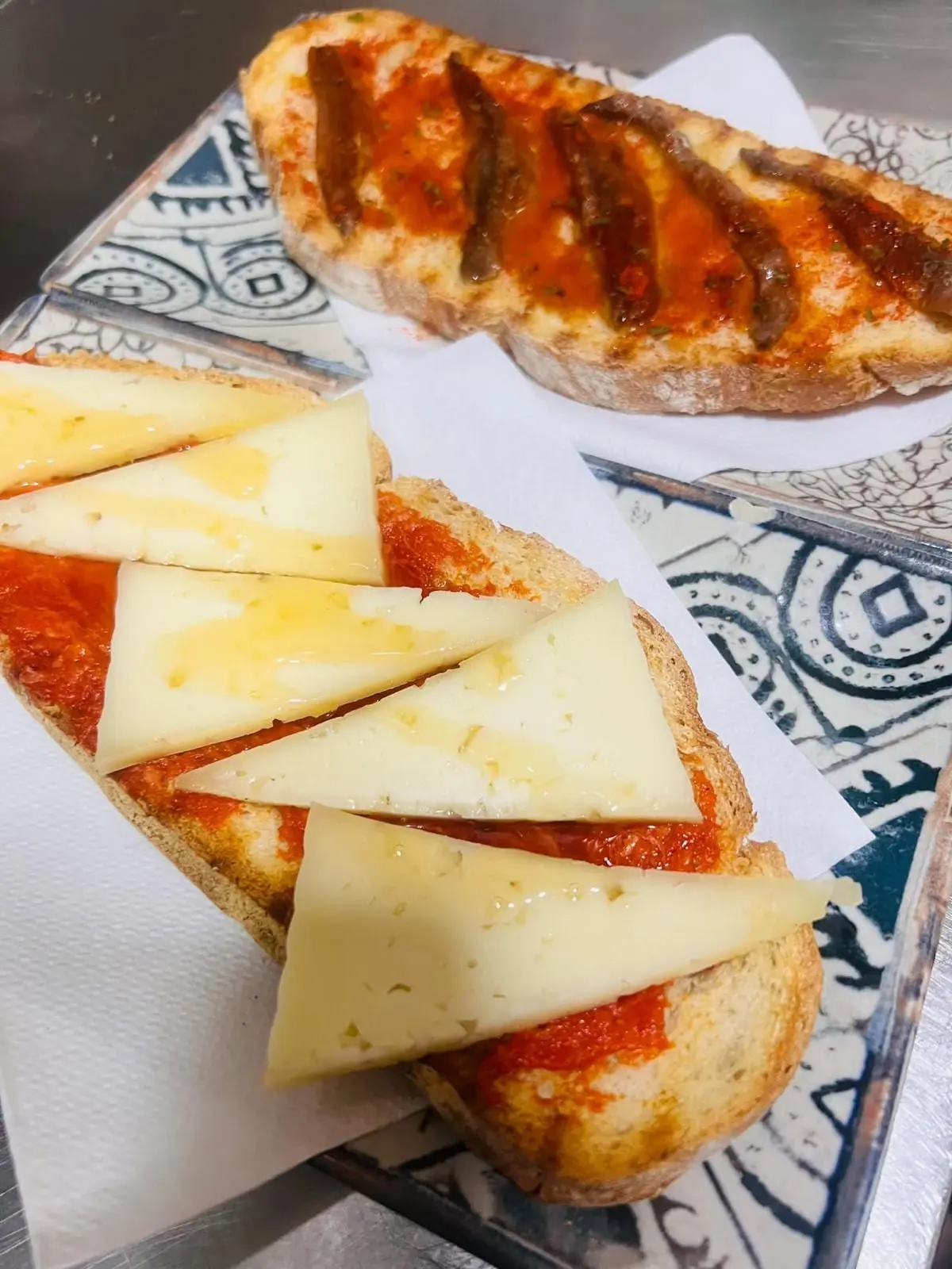 Dos torraetas servidas sobre azulejo: una de queso curado con tomate y otra de anchoas con tomate y pimentón.